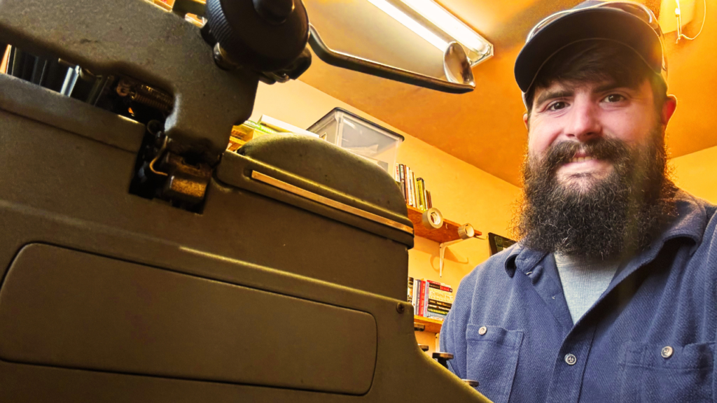 A man in a blue shirt sitting beside a typewriter.